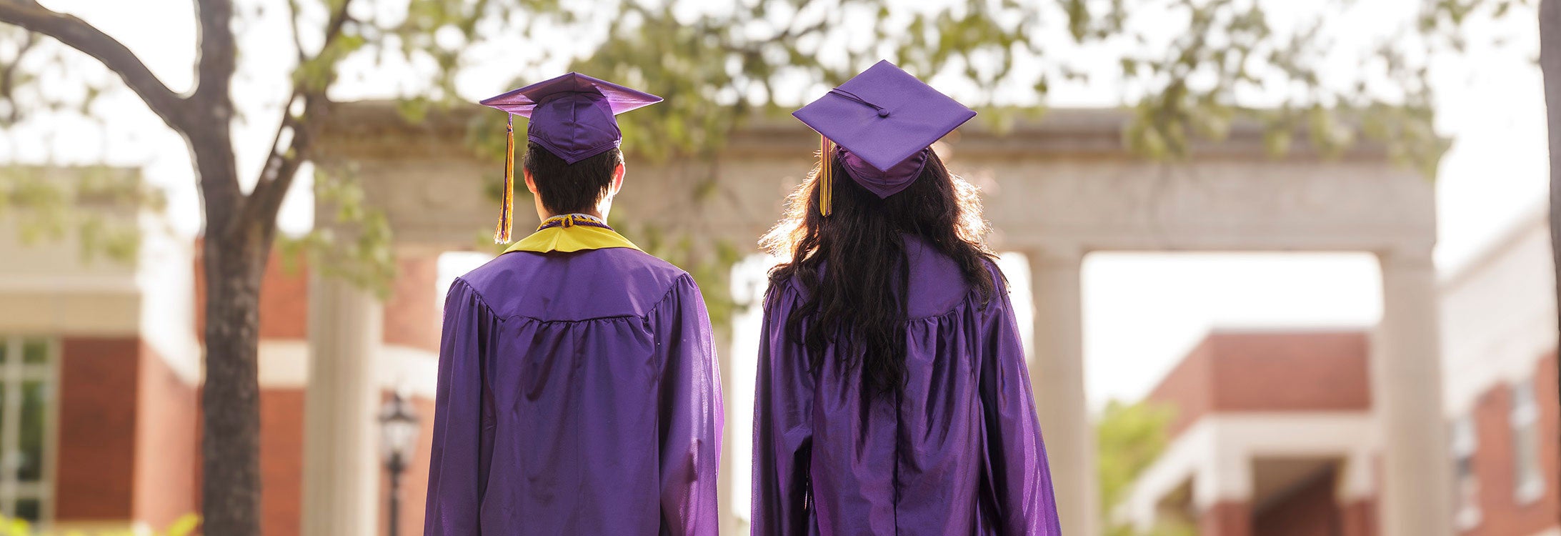 two students wearing a purple cap and gown facing away from the camera. University building in background.