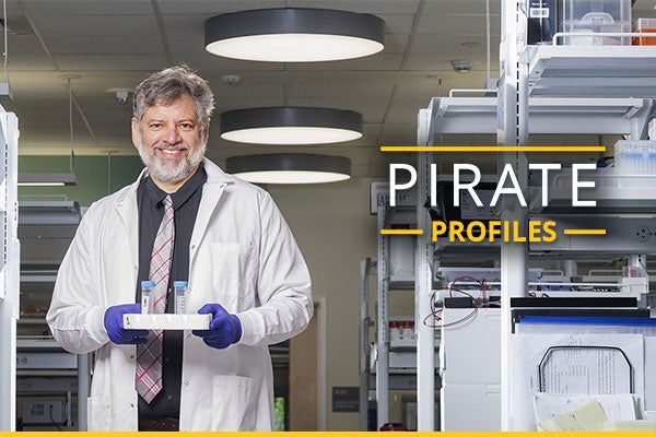 A man wearing a white coat and tie stands in a lab holding a tray of test tubes.
