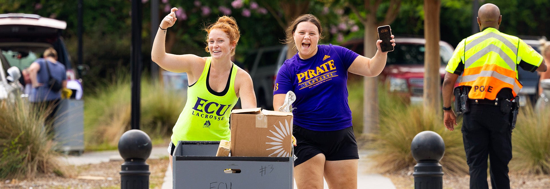 two female students in summer attire walk toward the camera holding up the pirate hook hand sign on move-in day.